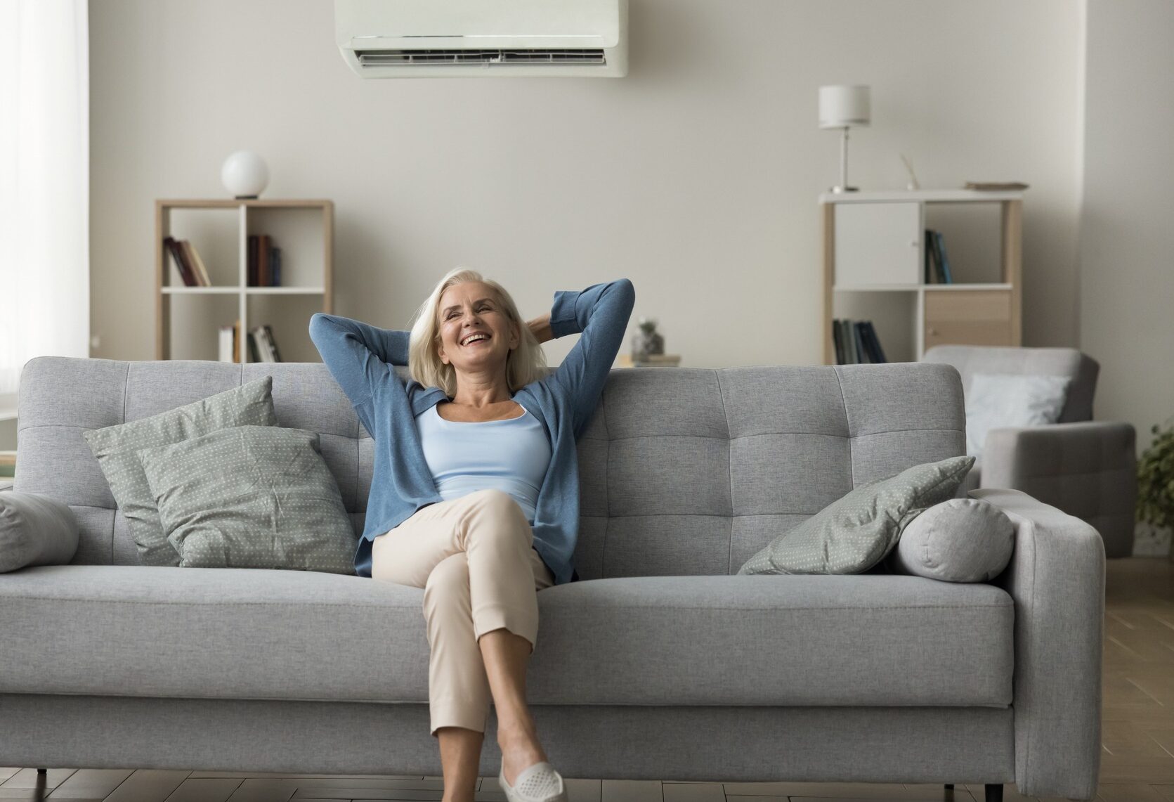 Cheerful old woman resting on grey soft couch under conditioner